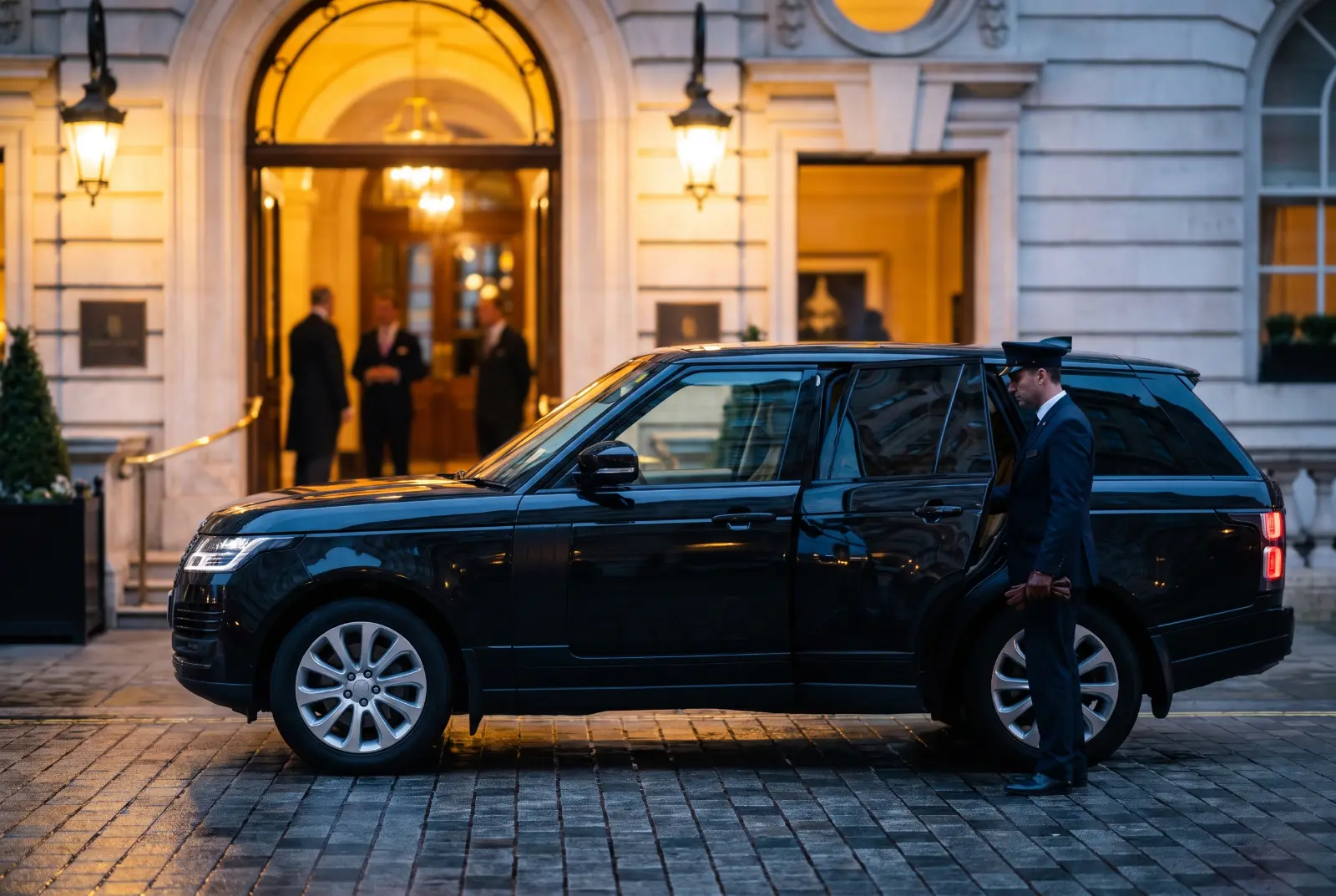 A chauffeur waiting by the open rear door of a black SUV outside a London hotel